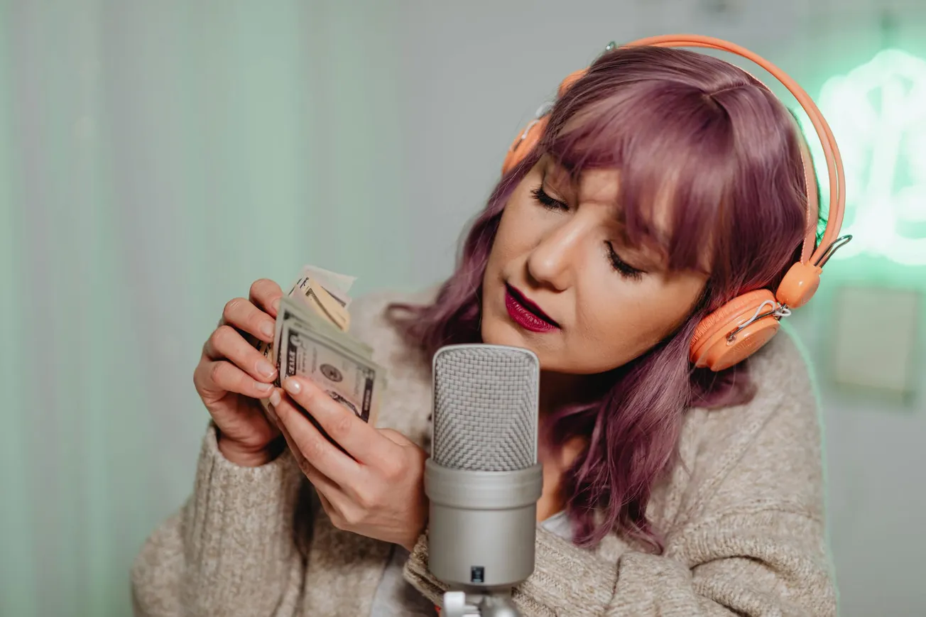 Woman with purple hair and orange headphones records ASMR video, softly counting dollar bills into a microphone.
