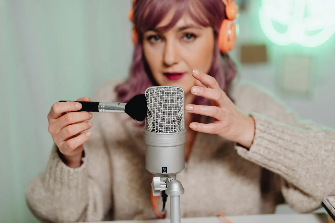 A woman with purple hair and orange headphones uses a brush near a microphone, creating an ASMR video. She appears focused and calm.