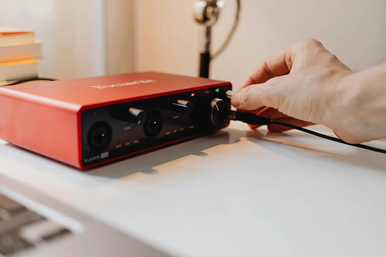 A hand adjusts a knob on a red audio interface on a desk. The device, with visible ports and lights, suggests a focused, tech-related setting.