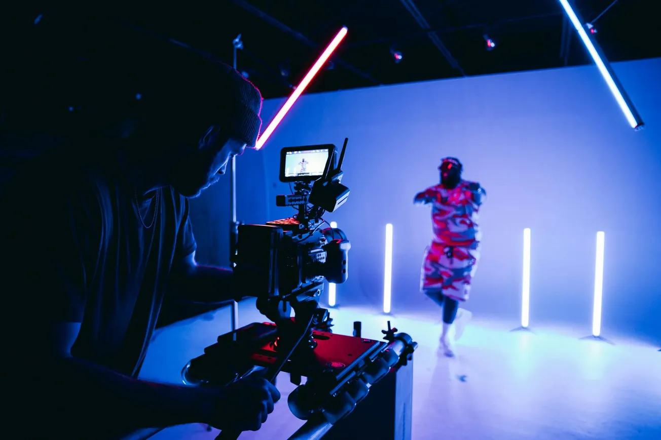 A person films a dancer in a dimly lit studio with vibrant neon lights, creating an energetic, futuristic vibe. The scene is dynamic and creative.
