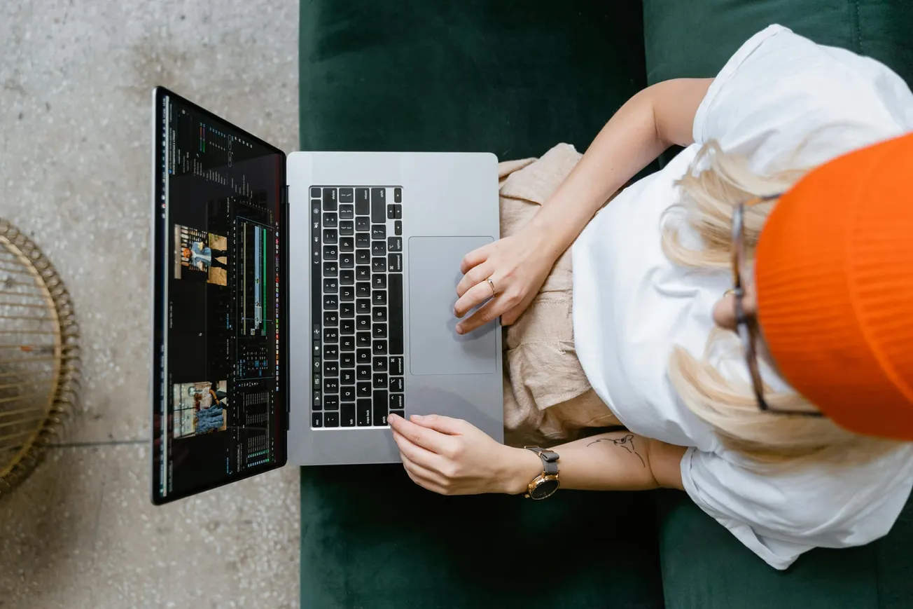 Overhead view of a person in an orange beanie editing video on a laptop. They sit on a dark green couch, conveying focus and creativity.