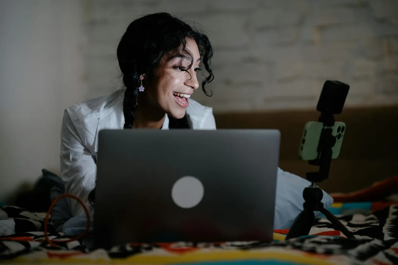 Smiling woman in a bright room, engaged in a video call on her laptop. A smartphone on a tripod is visible.