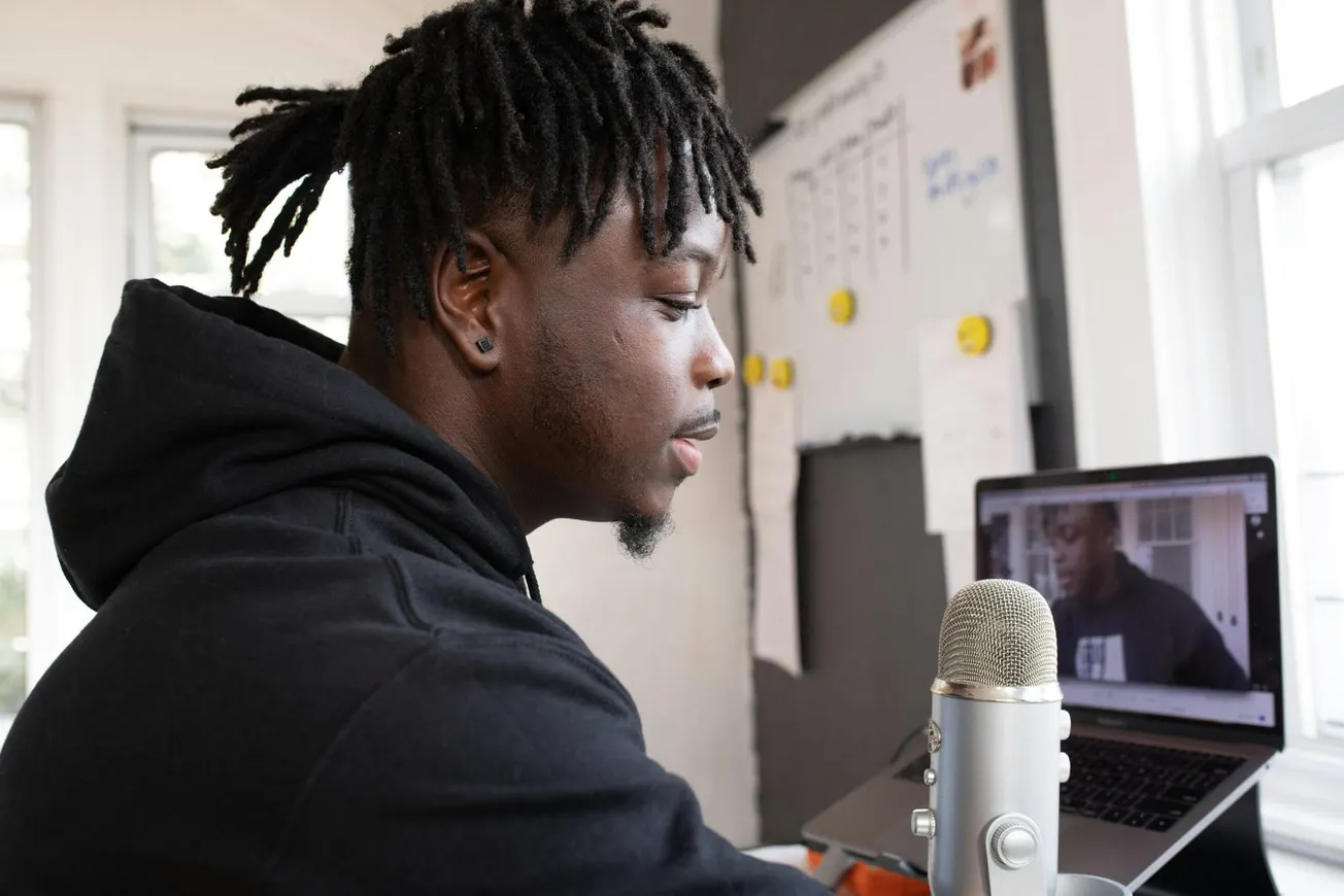 A person with dreadlocks wearing a black hoodie speaks into a microphone during a video call on a laptop. The scene is set in a bright room.