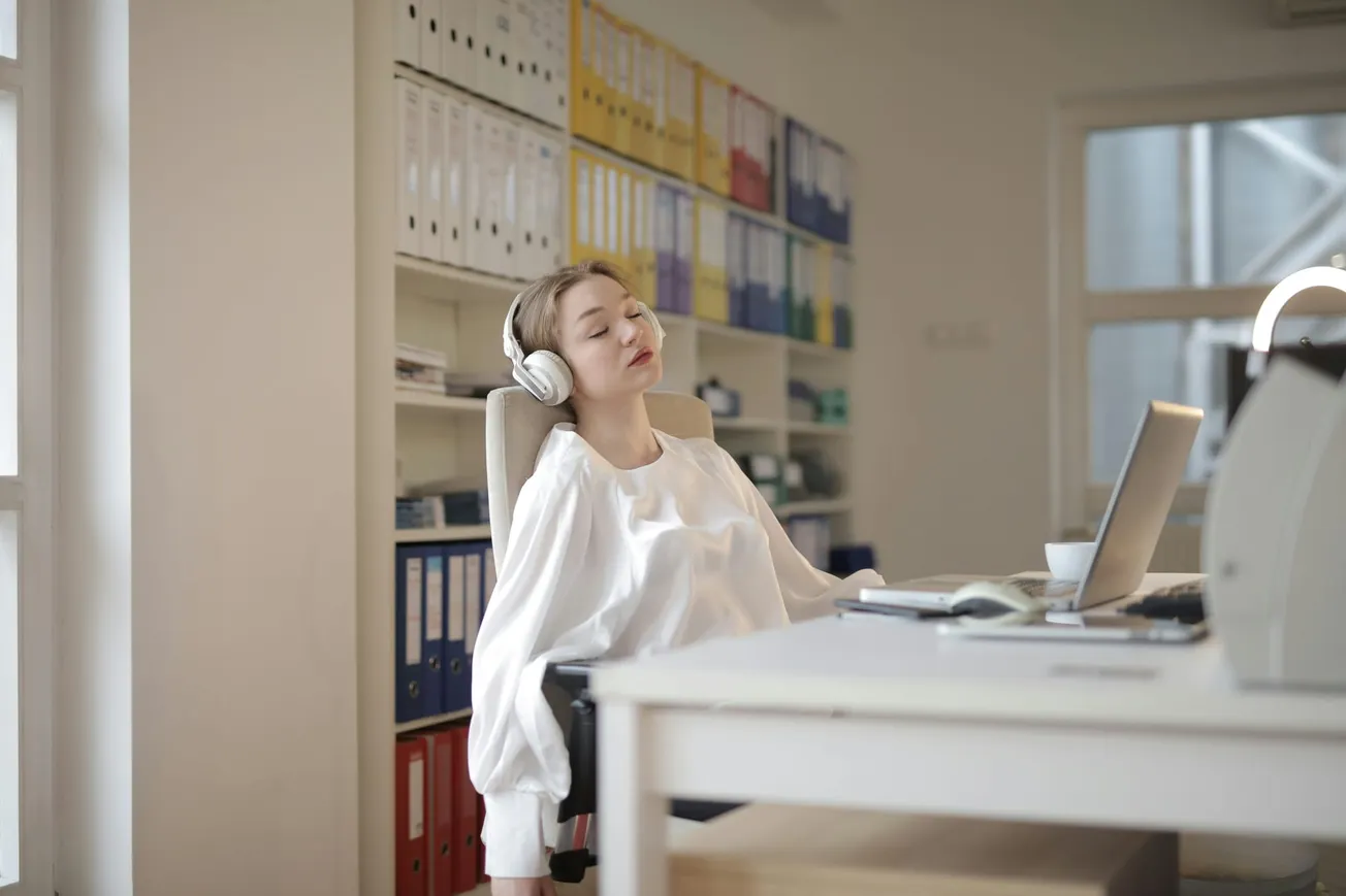 A woman in a white blouse relaxes with closed eyes and headphones at an office desk, surrounded by shelves of colorful binders.