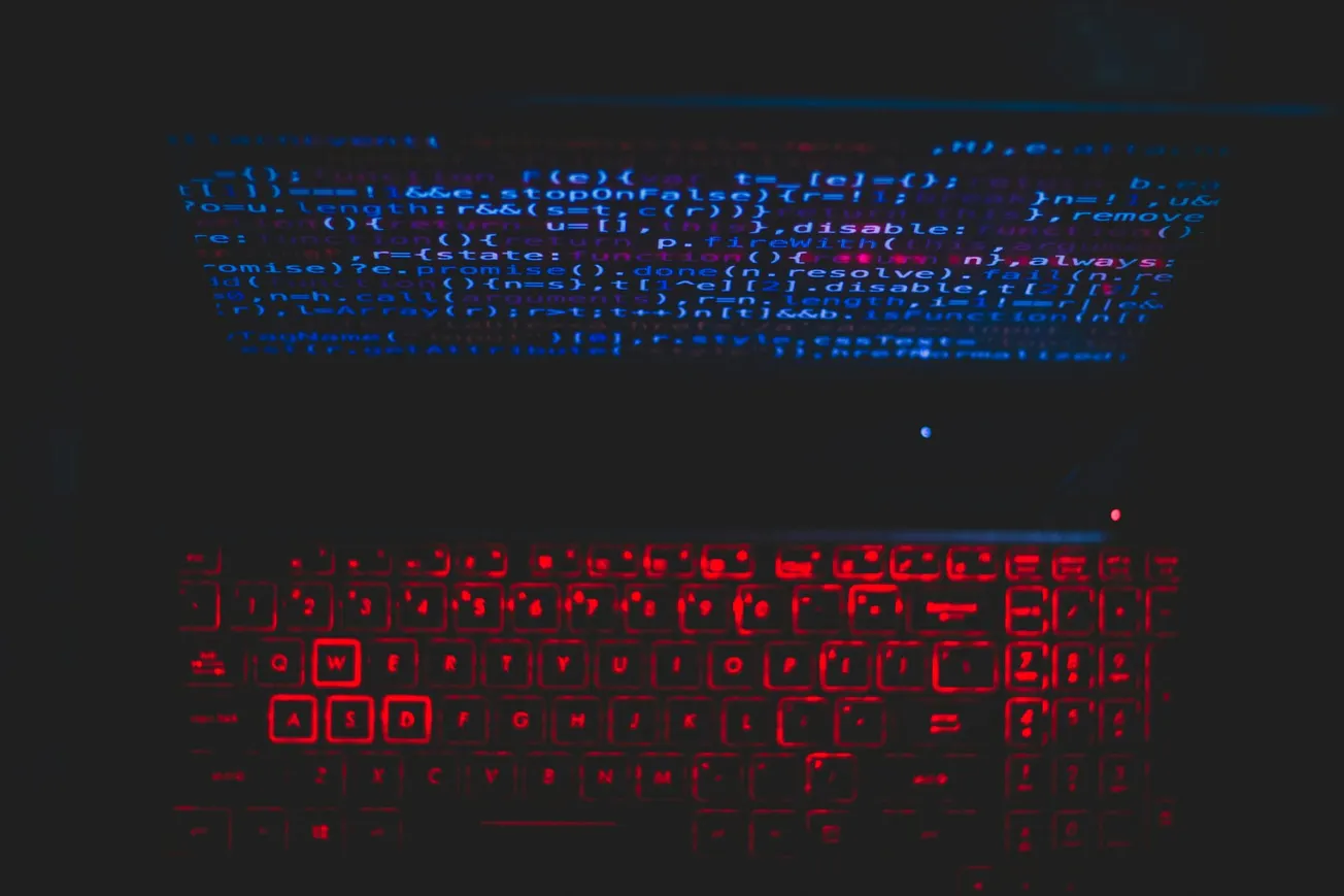 A laptop with red backlit keys shows blue code on its screen in a dark room, evoking a mysterious and high-tech atmosphere.