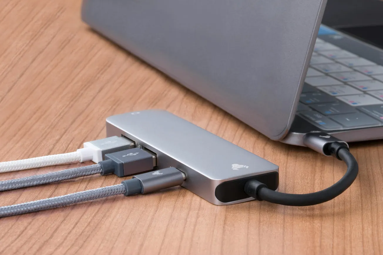 A sleek silver laptop on a wooden desk, connected to a silver multi-port USB hub with black, white, and gray cables. Modern, tech-focused ambiance.