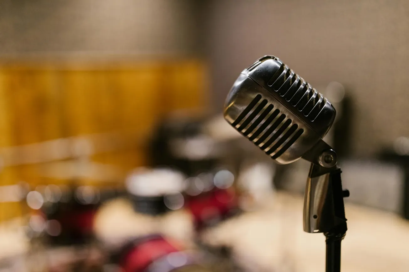 A vintage microphone is prominently displayed in a dimly lit recording studio, with blurred drum sets in the background.