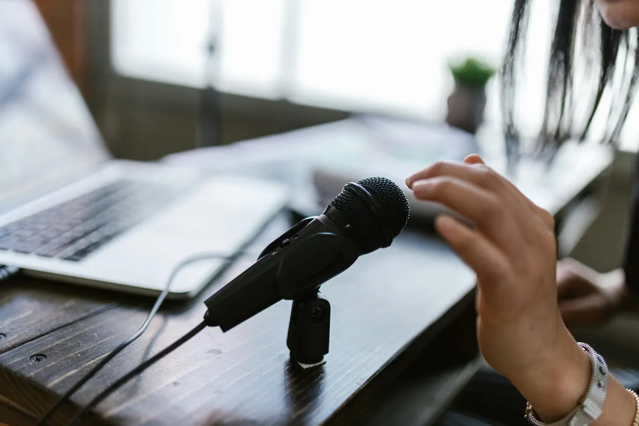 A person adjusts a microphone on a wooden desk beside an open laptop. The image conveys a focused and professional atmosphere, ideal for recording.