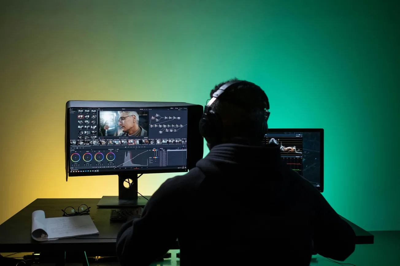 A person with headphones edits video footage on two monitors in a dimly lit room with green and yellow lighting. A notepad and glasses are on the desk.