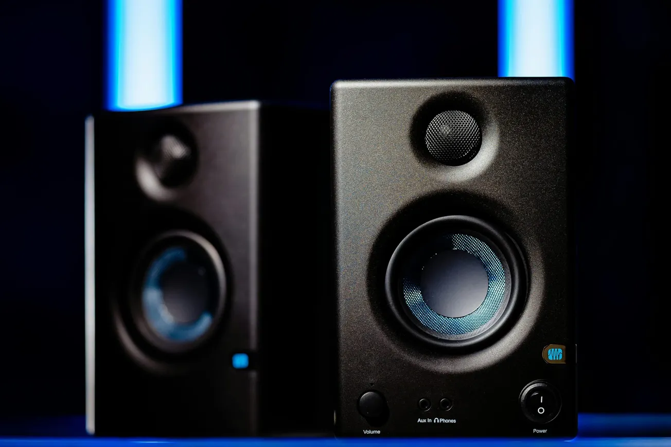 Close-up of two black speakers with blue accents, positioned side by side. Bright blue light bars in the background add a modern, sleek ambiance.