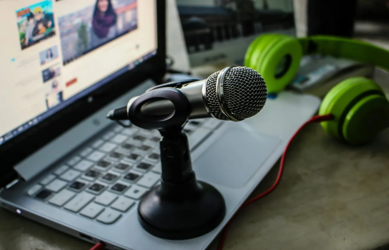 A microphone on a stand is placed on an open laptop displaying a video. Green headphones lie nearby. The setup suggests a casual podcast or recording session.