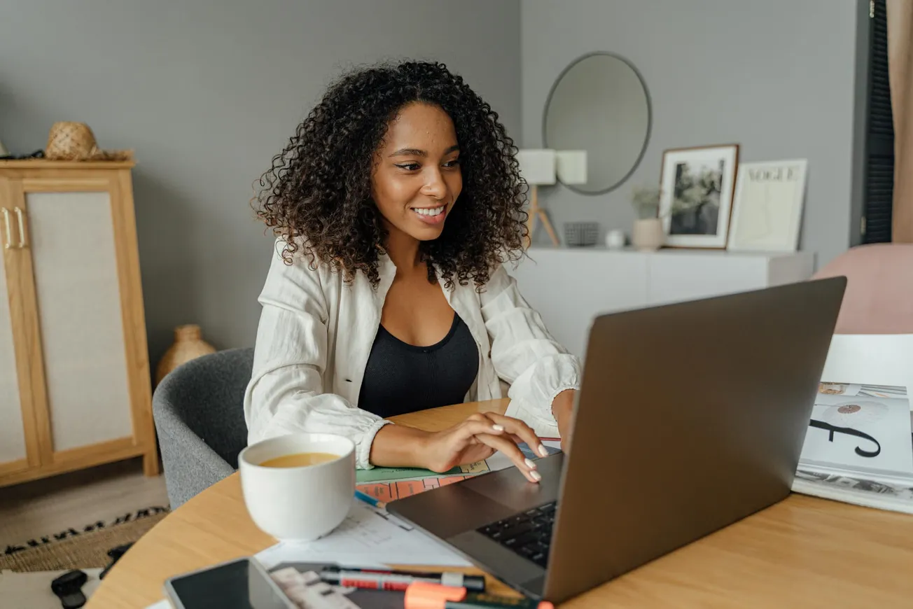 Smiling woman with curly hair works on a laptop at a wooden table, surrounded by notebooks and coffee.