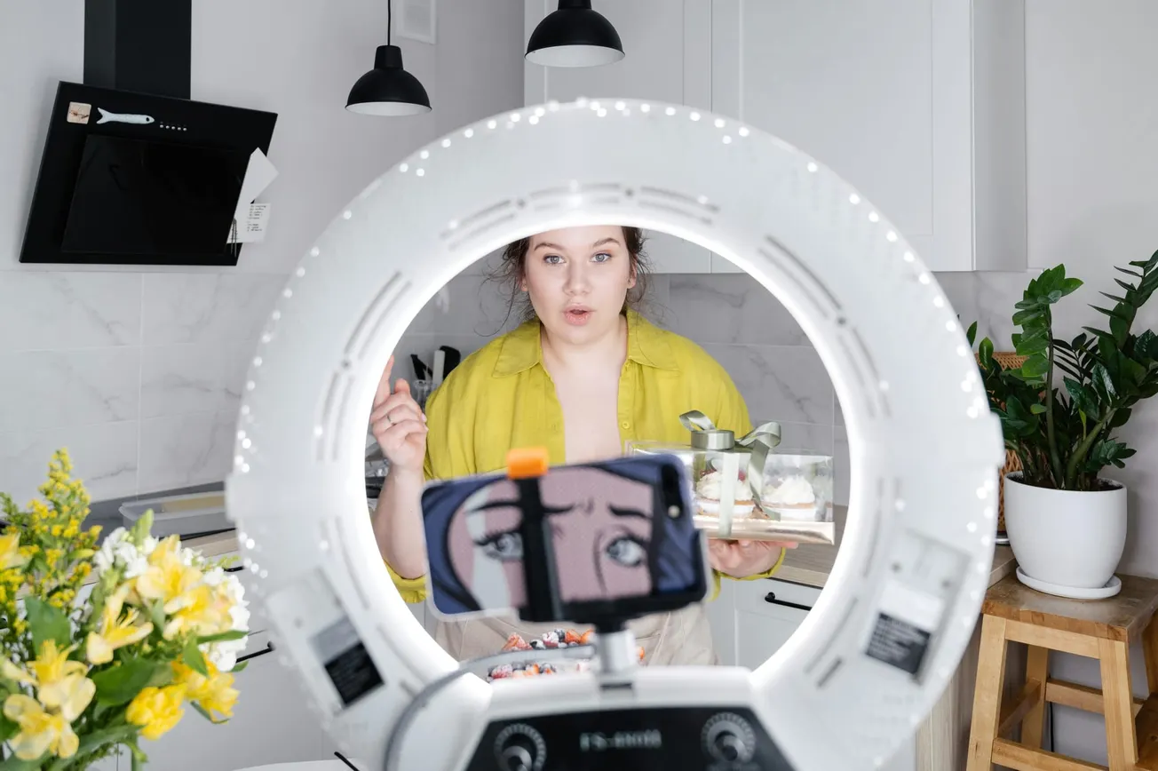 A woman in a kitchen, holding a tray of desserts, stands behind a ring light with a smartphone mounted. The setting is bright and lively.