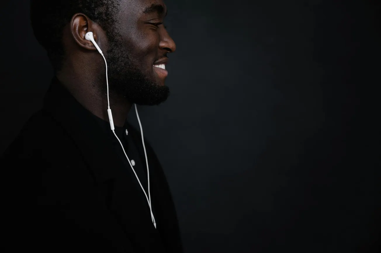 A man with earbuds smiles in profile against a dark background. He's wearing a black jacket, conveying a relaxed, joyful mood.