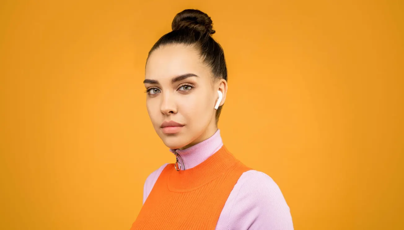 Woman with hair in bun, wearing AirPods, an orange sleeveless top over a lavender turtleneck, against an orange background, looking at the camera.