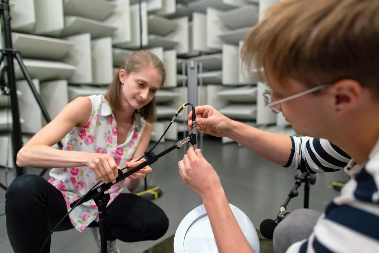 Two engineers adjusting audio equipment and microphone setup inside an anechoic soundproof testing chamber.