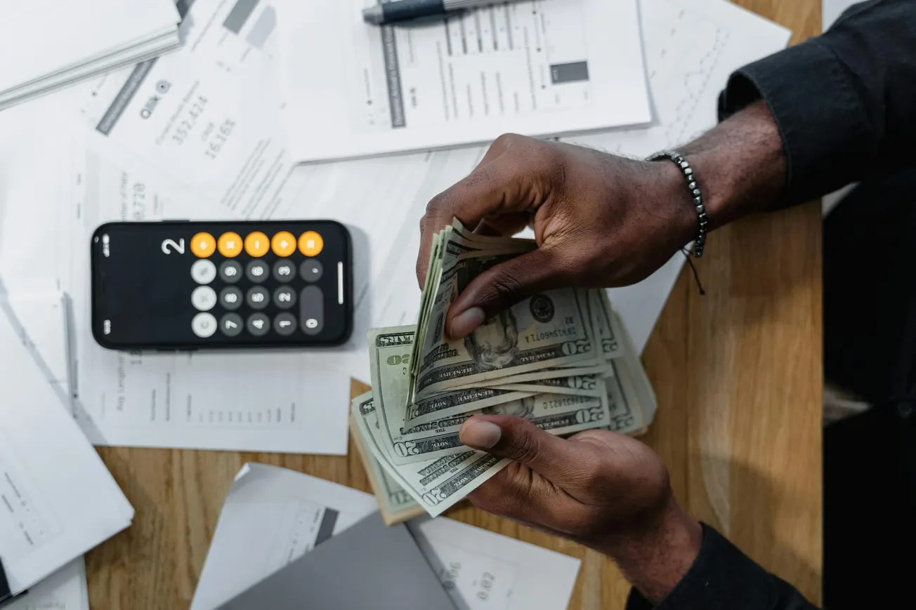Hands counting dollar bills over a cluttered table with scattered papers, a smartphone calculator, and a pen, conveying a sense of financial management.