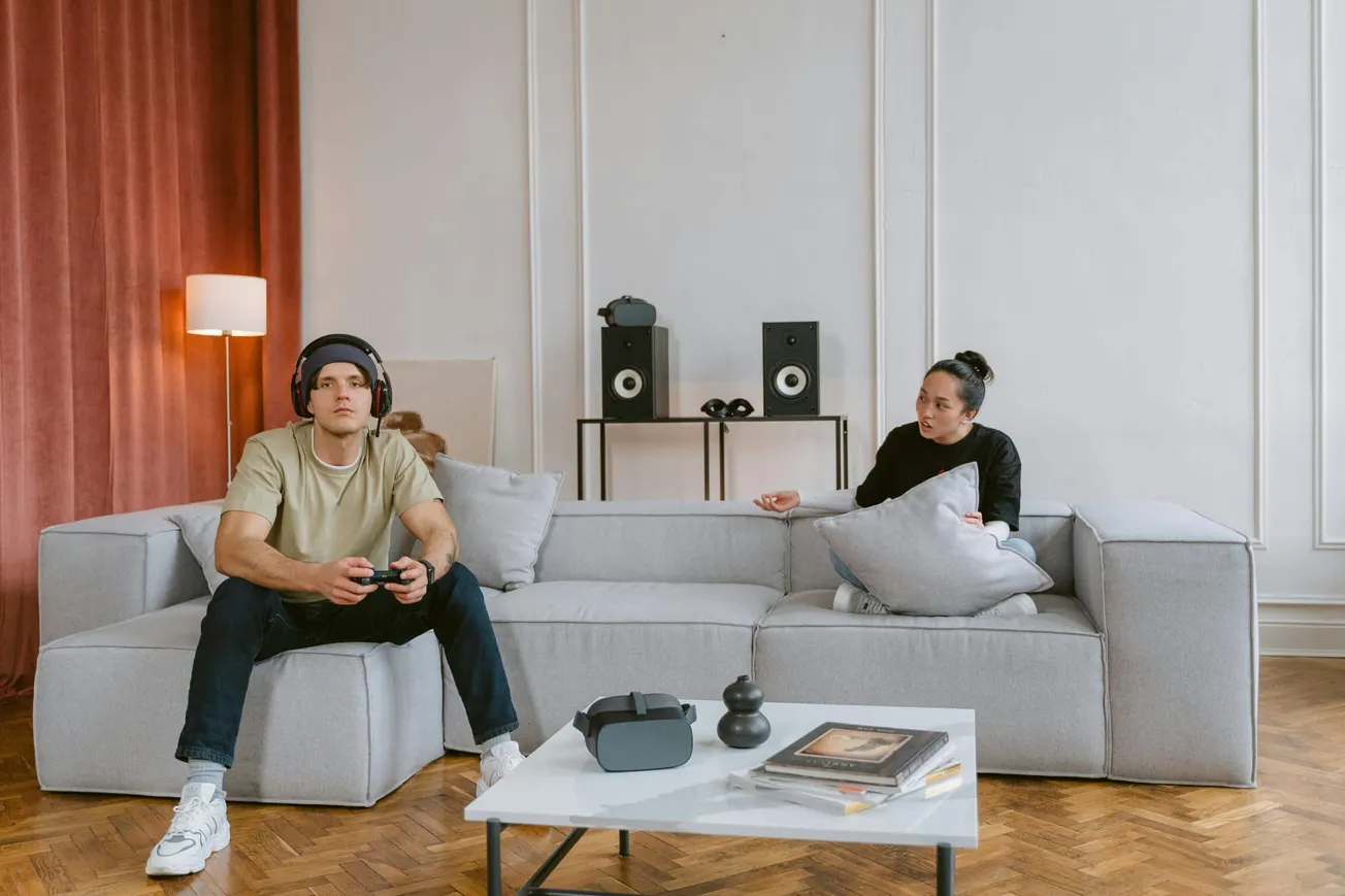 A man with headphones plays video games on a gray sofa, while a woman sits nearby holding a pillow. Speakers and decor create a cozy, relaxed atmosphere.