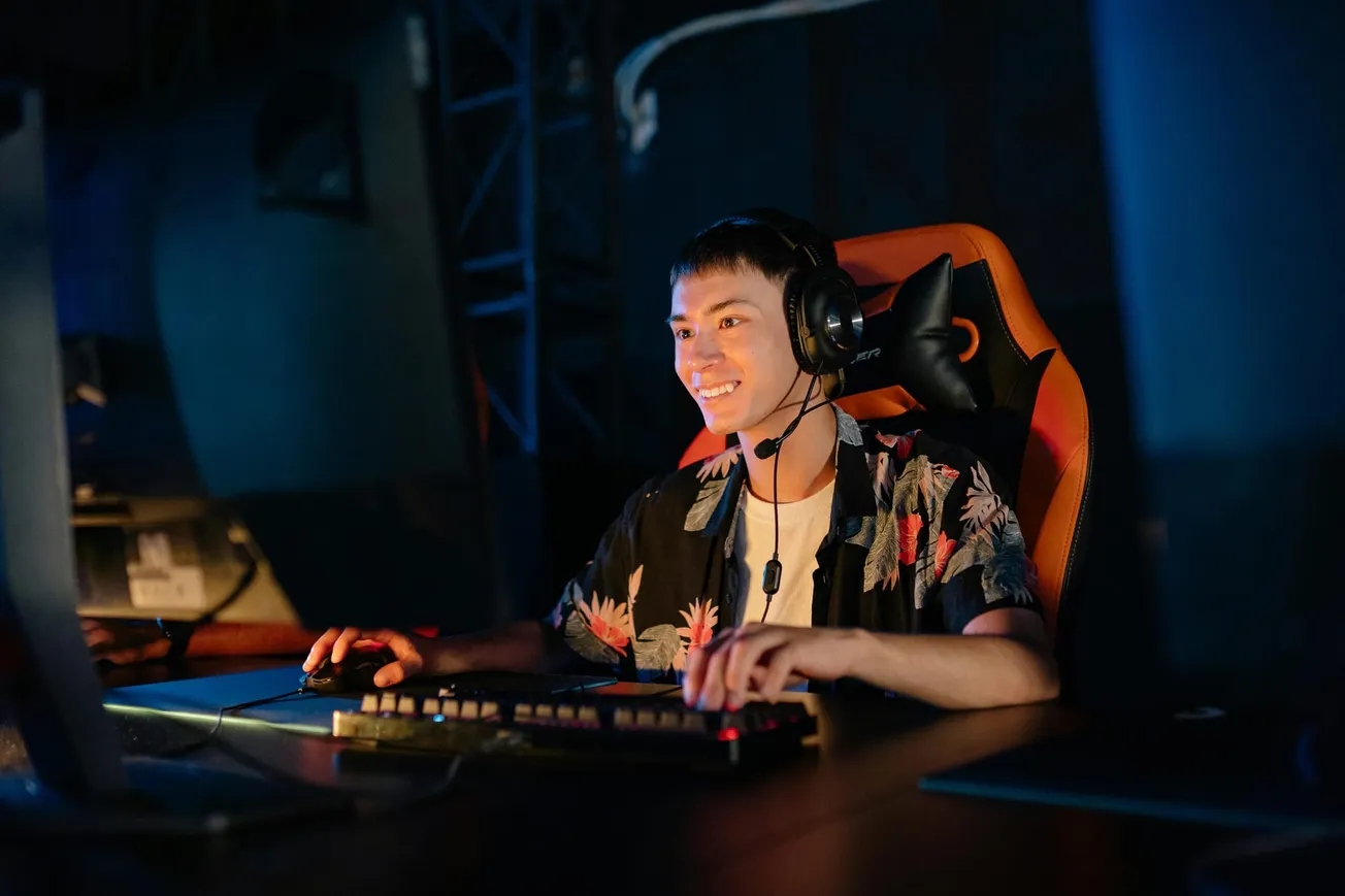 Young man gaming with a headset, smiling, seated in an orange gaming chair. He wears a floral shirt, with a relaxed and focused expression in dim lighting.