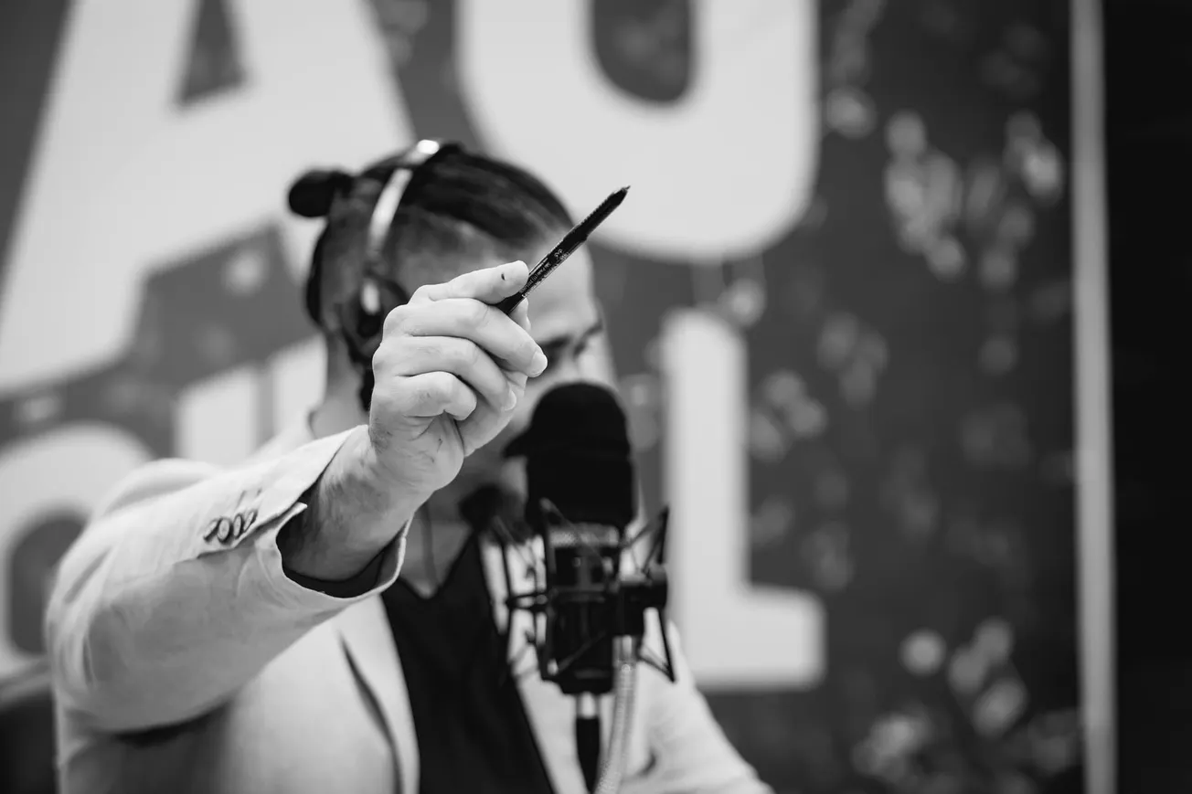 A man holds a pen in front of a microphone, face obscured, conveying focus and concentration. The background features bold, blurred text. Black and white image.