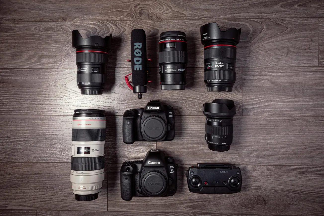A collection of photography gear on a wooden floor, including six camera lenses, two Canon DSLRs, a Rode microphone, and a drone controller.