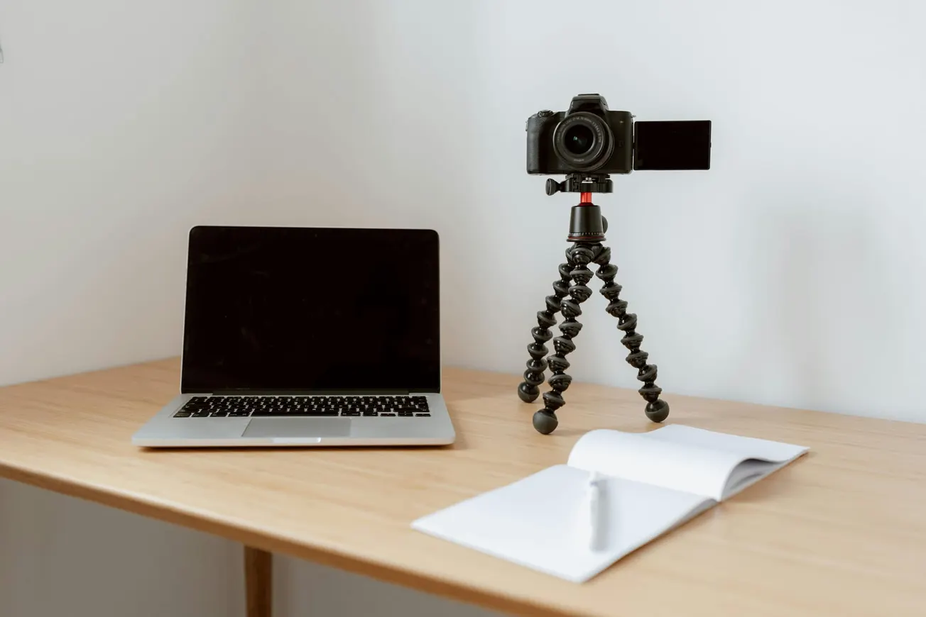 A laptop, camera on a flexible tripod, and an open notebook with a pen are on a wooden desk against a plain white wall, creating a clean, organized workspace.