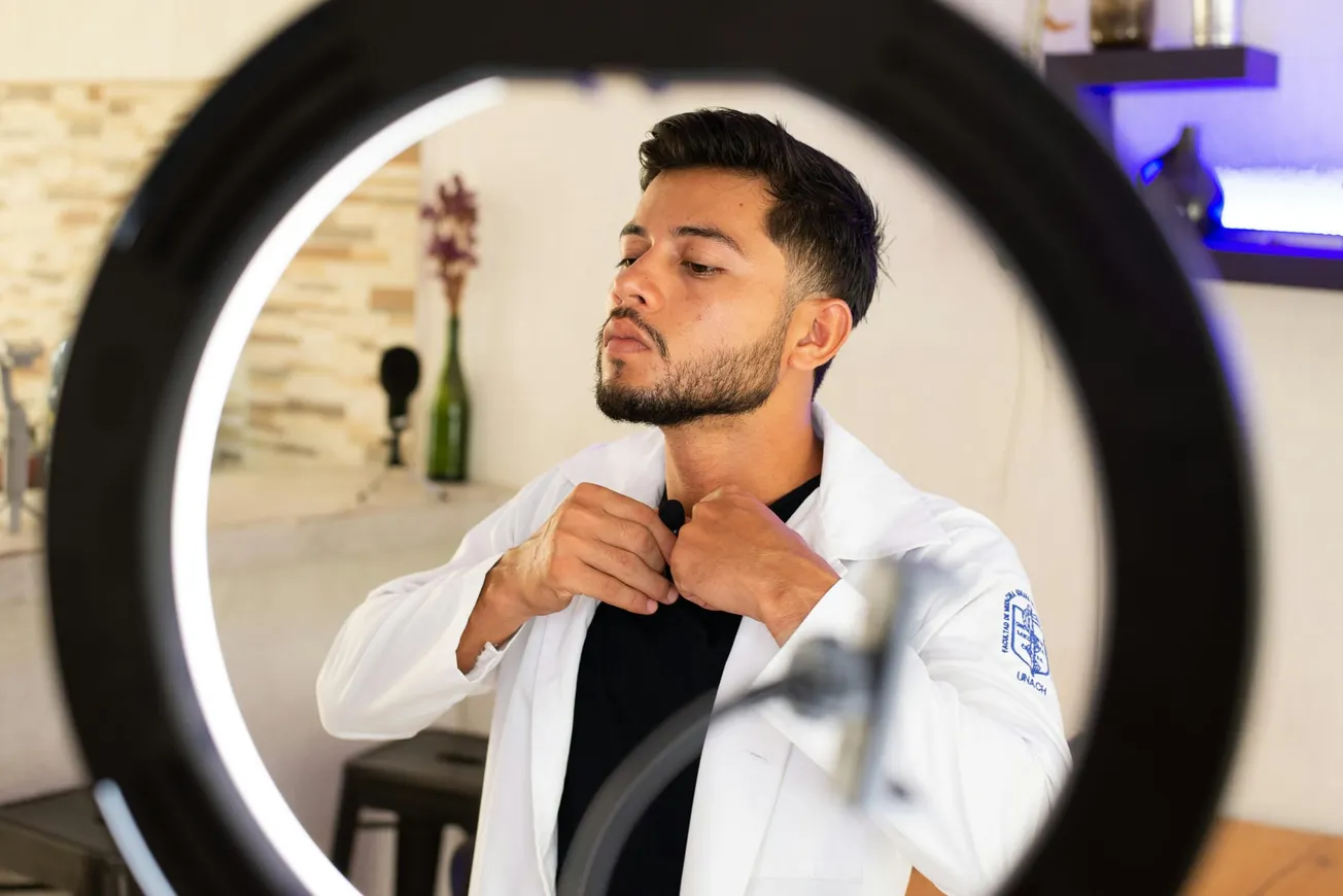 A man adjusts his shirt collar, captured through a ring light. He wears a white coat, suggesting a professional setting.