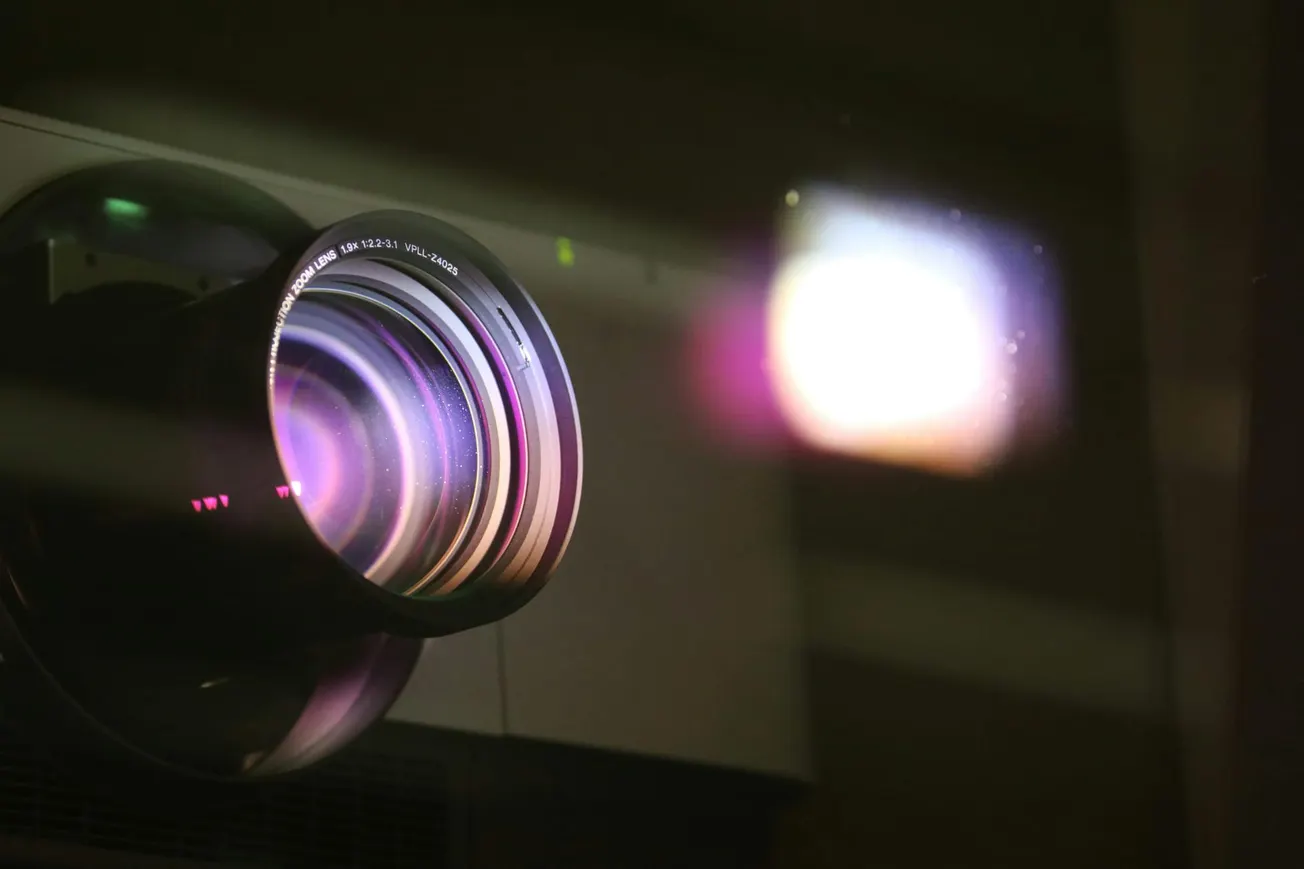 Close-up of a projector lens emitting a bright beam of light. The lens reflects purple and blue hues, creating a modern, technological feel.