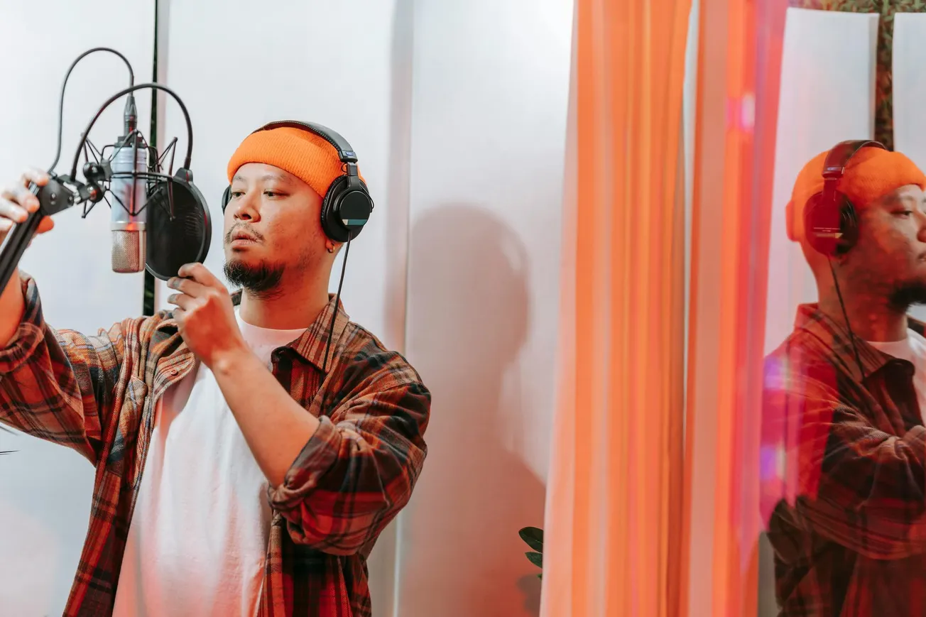 A man in an orange beanie and headphones adjusts a microphone in a recording studio. His reflection is visible in a nearby glass, creating a focused atmosphere.