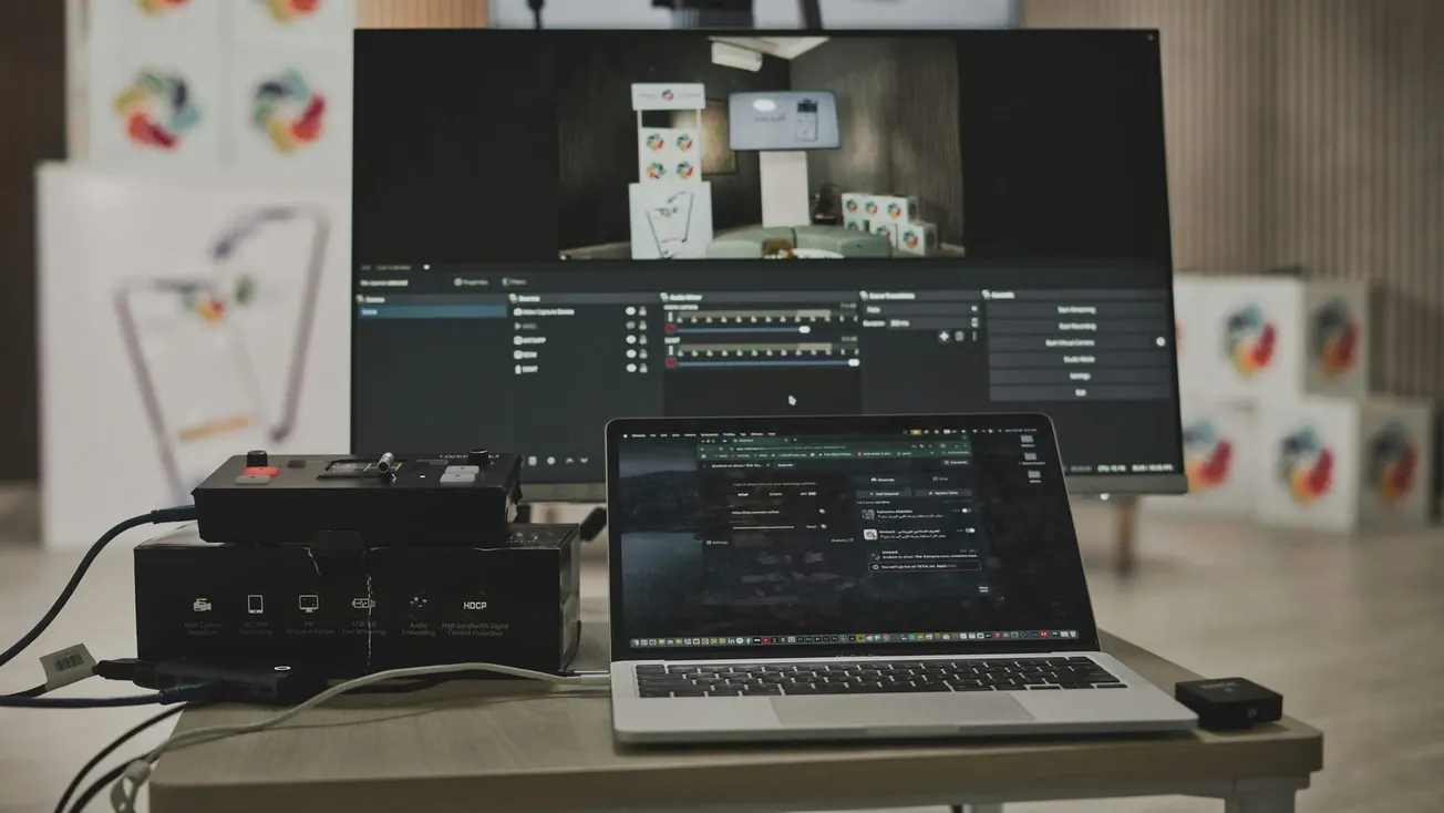 A laptop and electronic equipment on a desk in front of a large screen displaying a software interface. The scene has a tech-focused, professional tone.