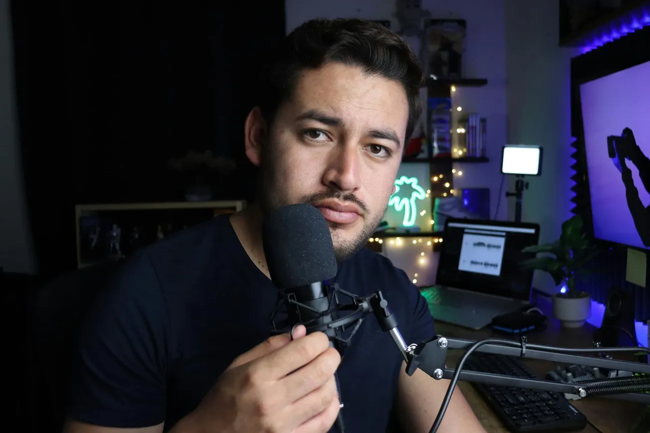 Young man with a serious expression holds a microphone in a dimly lit room. Background shows a laptop, neon light, camera gear, and blue accents.