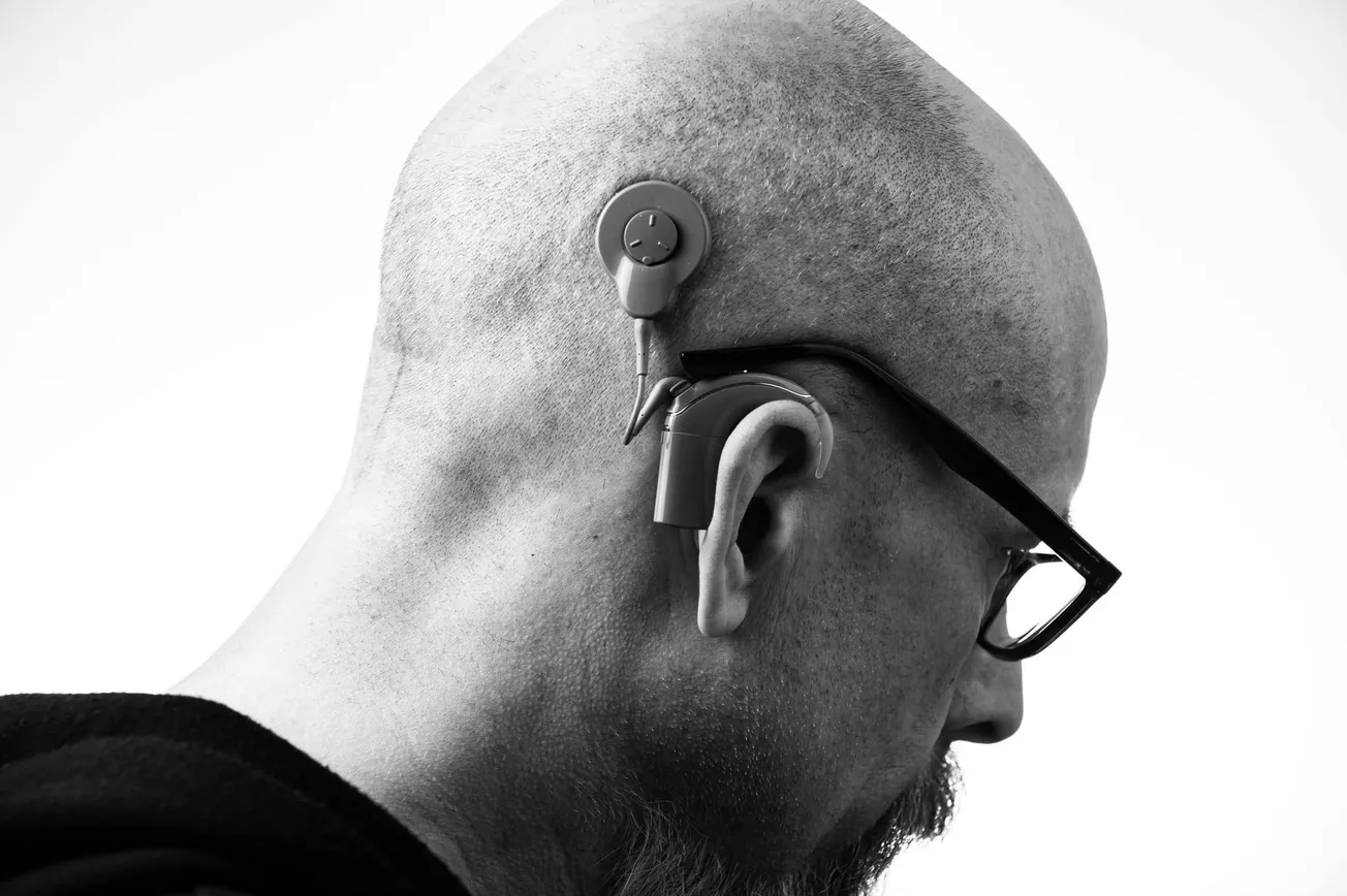 A bald man in profile wears glasses and a cochlear implant on his right ear, highlighting assistive technology. The mood is focused and contemplative.