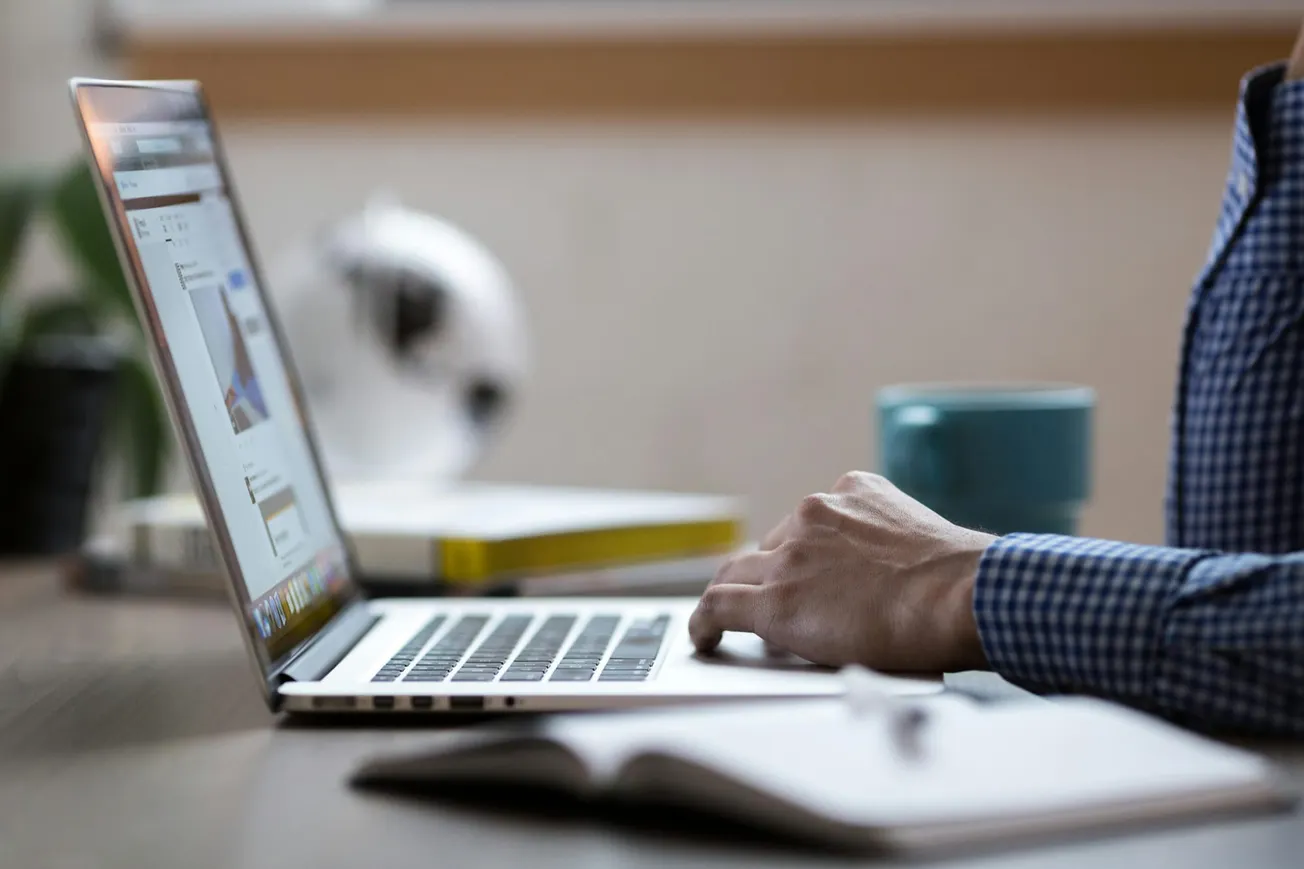 A person in a checkered shirt works on a laptop at a desk, with a notebook, book, and teal mug nearby. The scene conveys focus and productivity.
