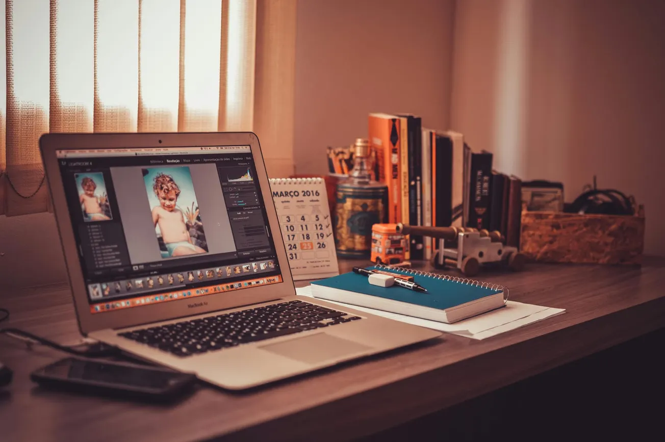 A cozy work desk with a laptop editing photo of a child, surrounded by books, a notebook with pens, and a March 2016 calendar.