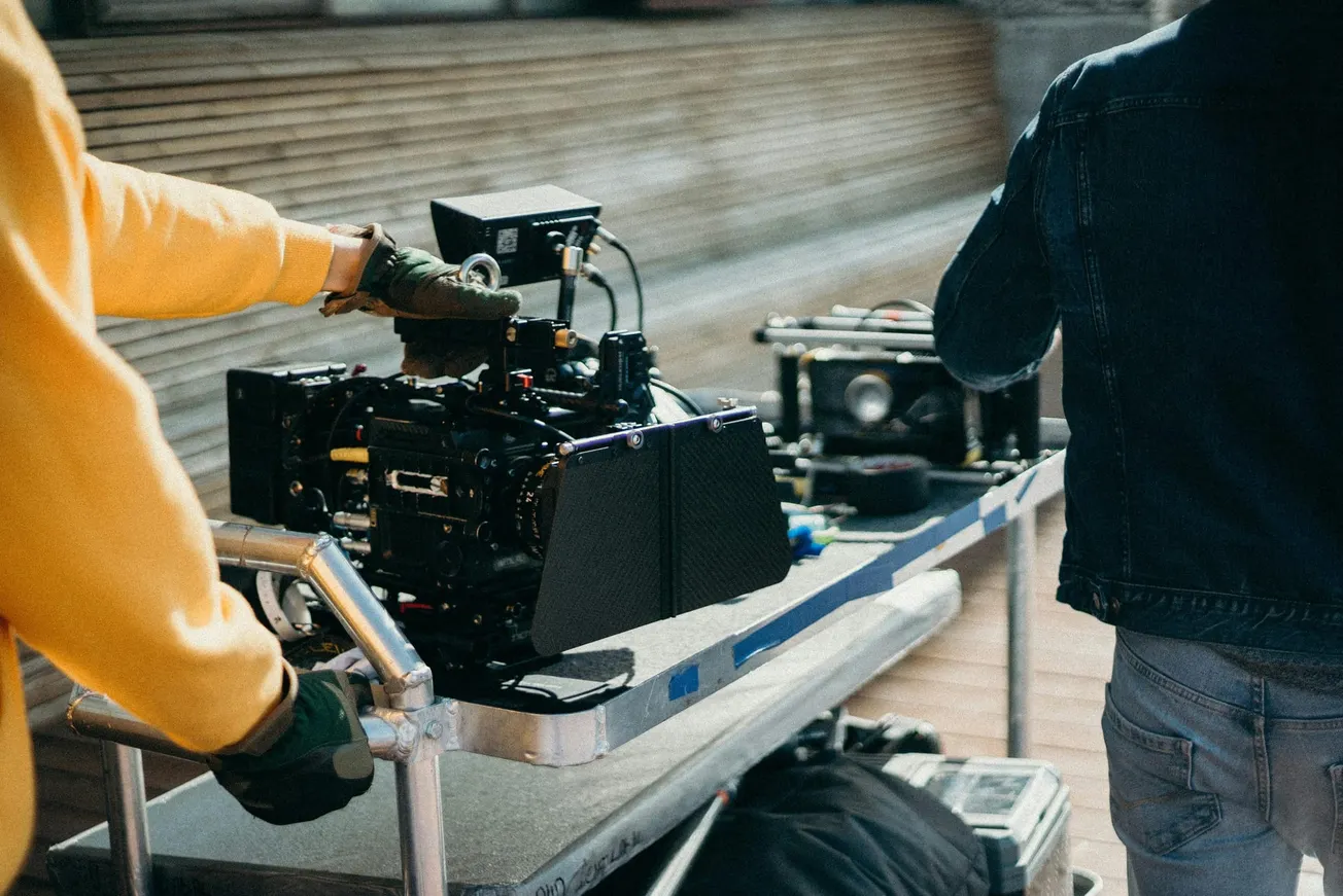 A person in a yellow sweater operates a film camera on a trolley. The scene conveys a focused, professional mood in a creative setting.