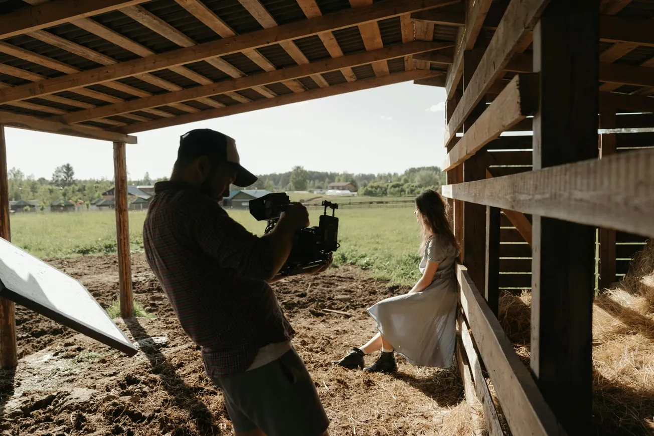 A filmmaker in a plaid shirt films a woman in a white dress sitting in a barn, with countryside and soft natural light creating a serene atmosphere.