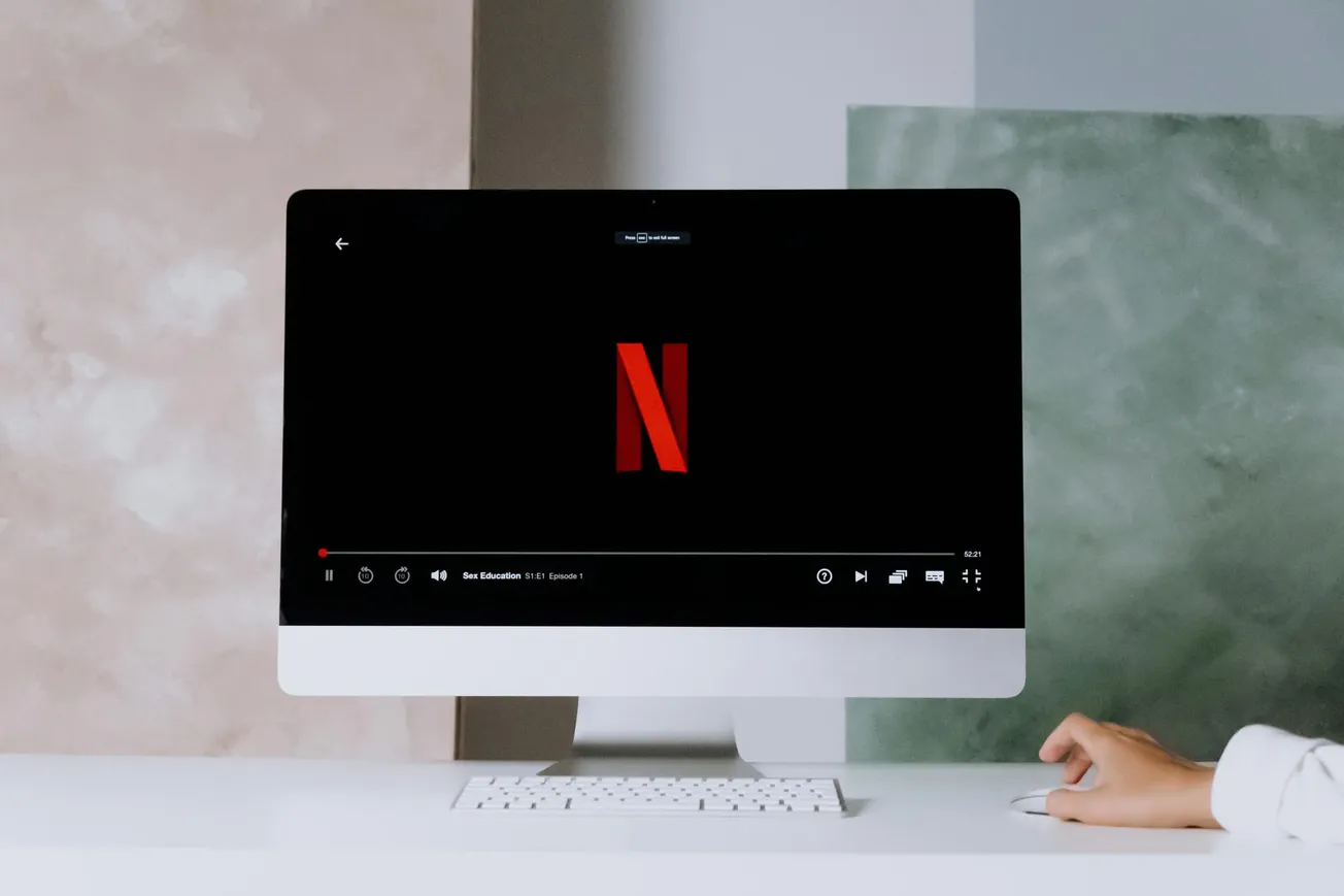 A computer monitor on a white desk displays the Netflix logo and a video progress bar. A hand is near a mouse, suggesting a calm, modern setup.