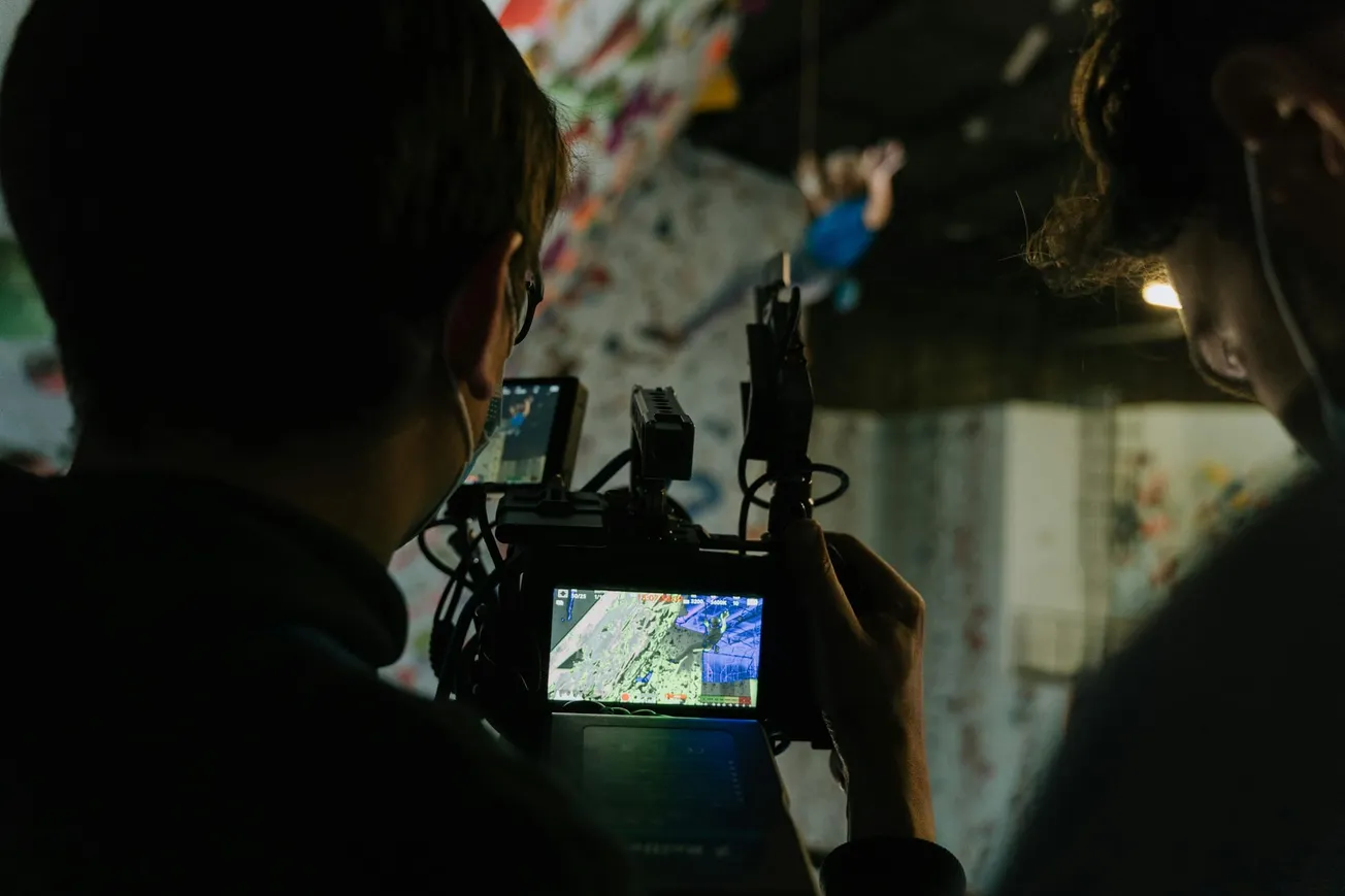 Two people filming a climber on an indoor rock wall. The camera screen shows the climber in focus. The setting is dynamic and action-oriented.