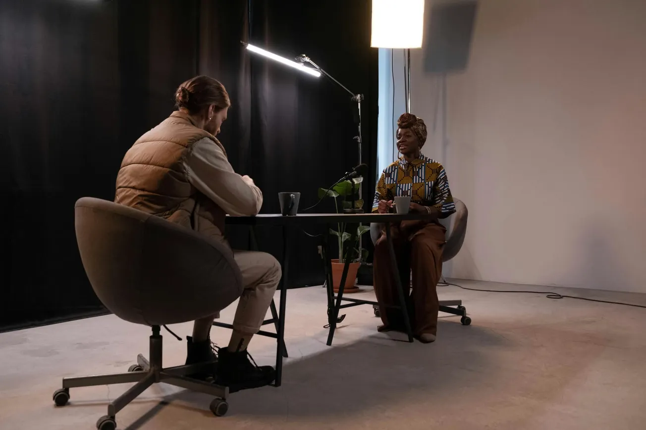 Two people sit across from each other at a table in a dimly lit room with a relaxed, cozy atmosphere. There is a potted plant nearby.