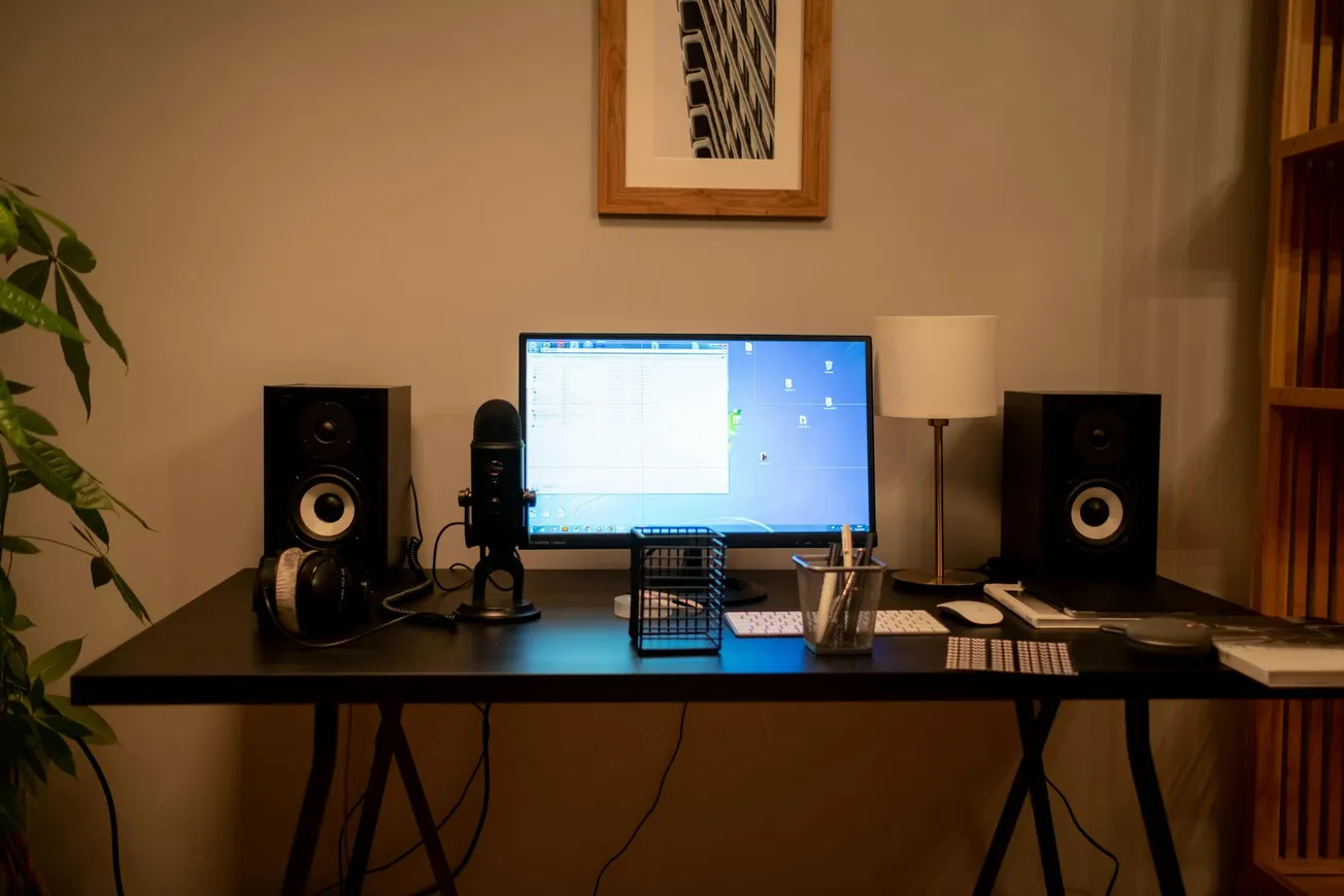 A tidy desk setup features a computer monitor between two speakers, a microphone, and headphones. A modern lamp and framed art add a cozy touch.