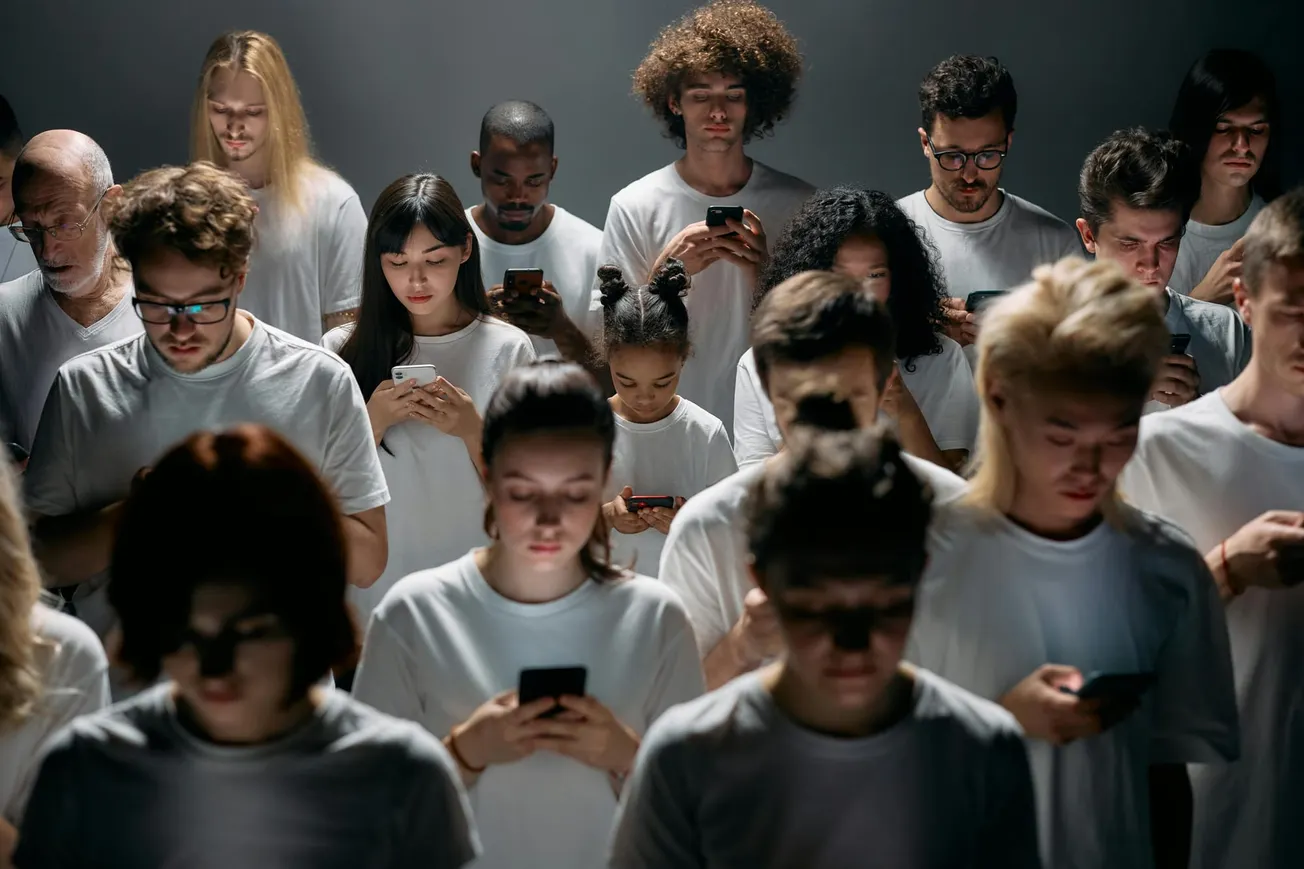 A diverse group of people in white shirts stands closely together, all looking down at their smartphones, creating a sense of disconnection and focus.