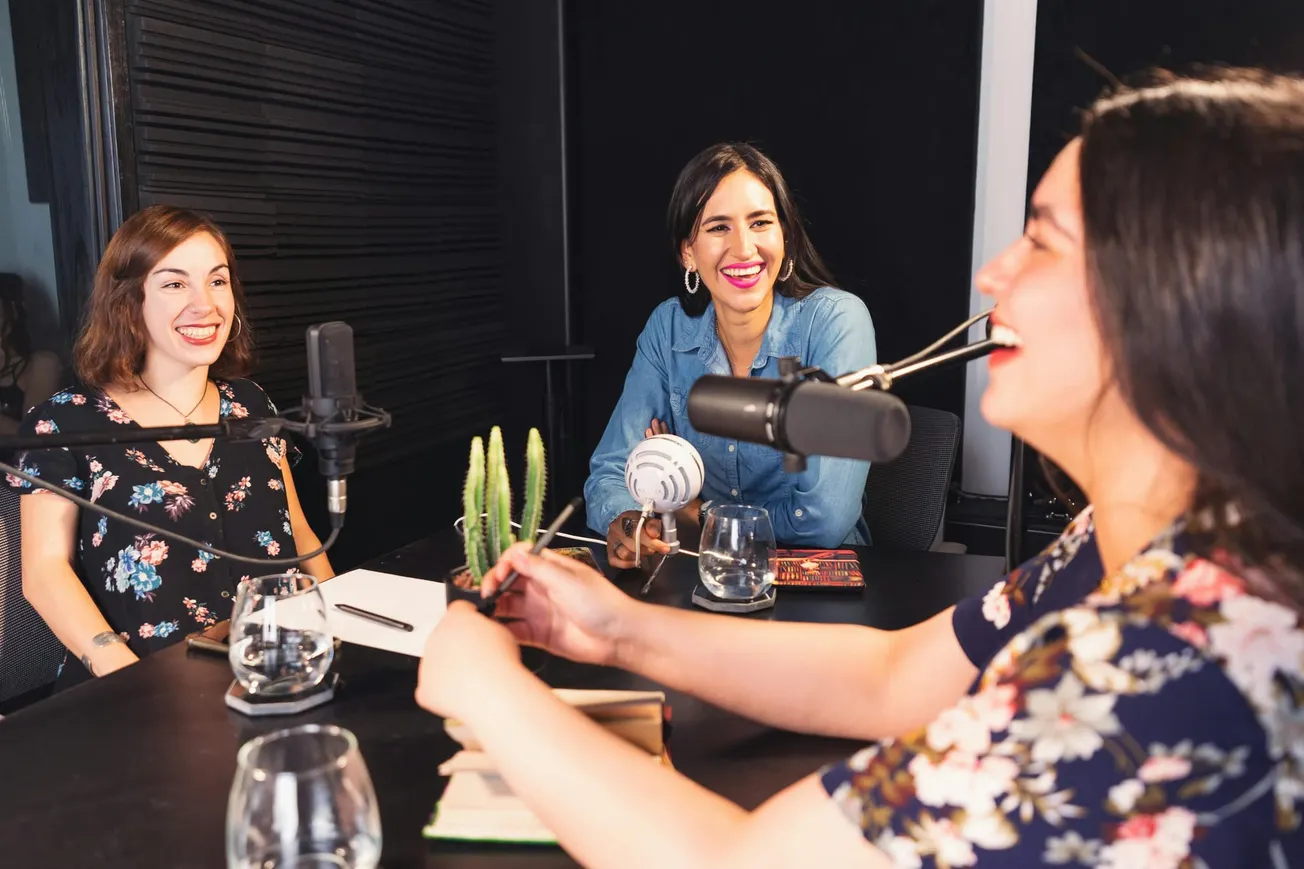 Three women sit around a table with microphones, laughing and recording a podcast.
