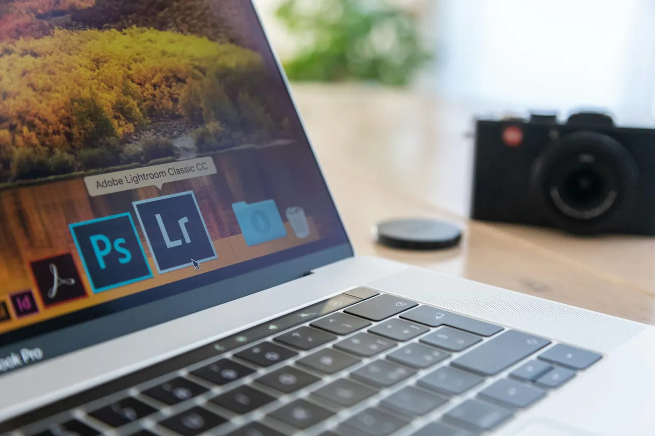 A laptop displays Adobe Photoshop and Lightroom icons on its screen. A camera and lens cap sit blurred in the background, suggesting a photography theme.