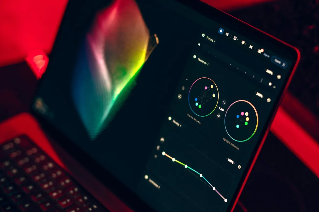 A laptop display shows colorful video editing software with color wheels and an adjustment graph. The ambient lighting is a warm red, creating a focused atmosphere.