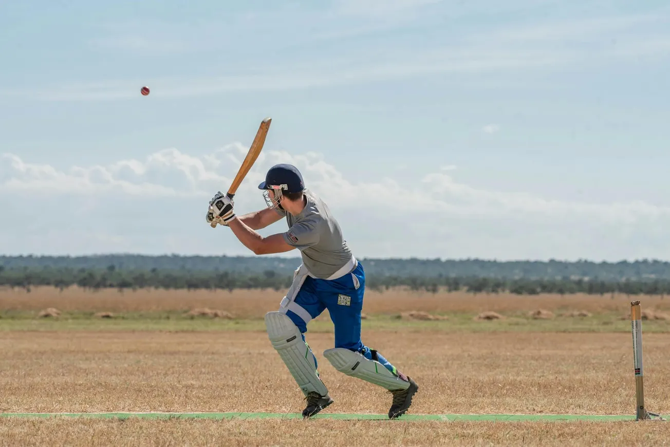 A cricket player in gear, poised to hit a ball mid-air on a sunny field.