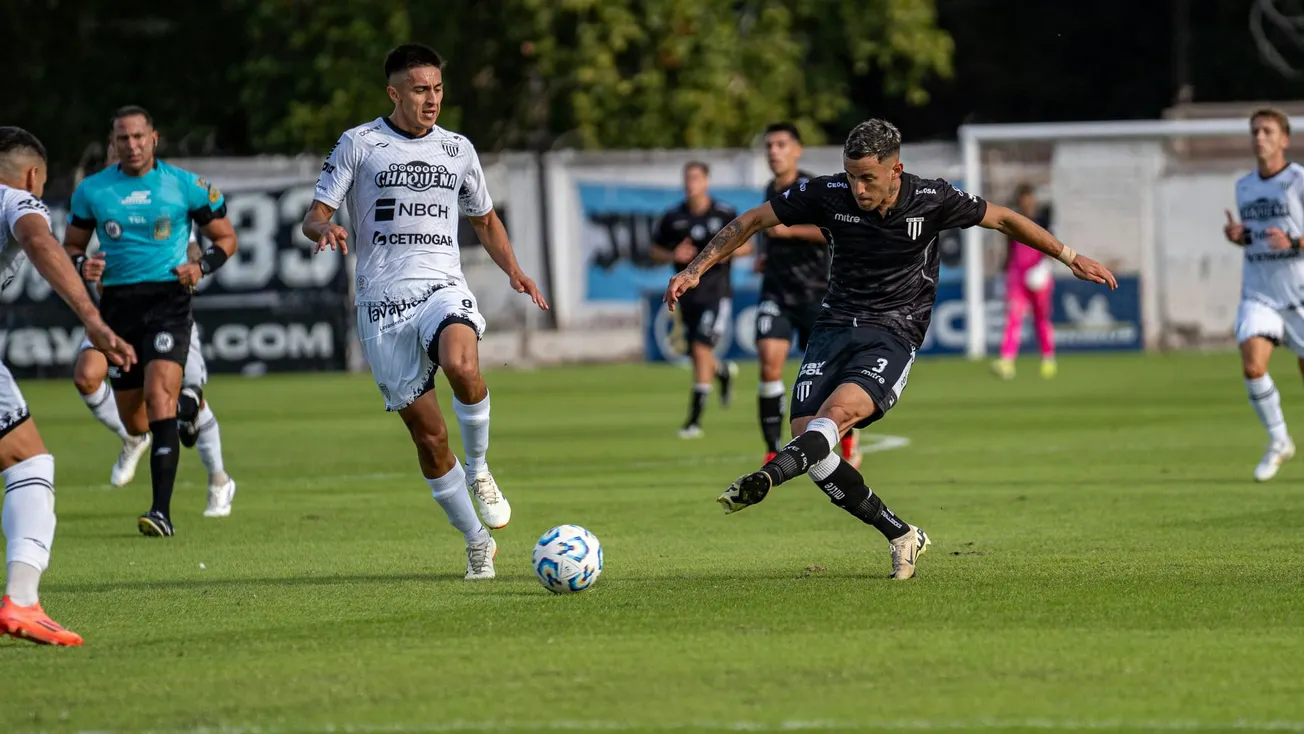 A soccer player in a black jersey fiercely kicks a ball on a green field, chased by another in white. Teammates and referee watch intensely, conveying action and competition.
