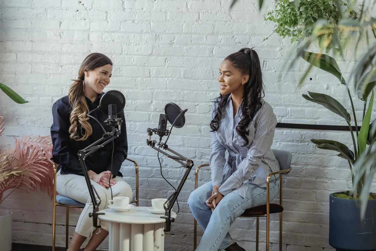 Two women sit in a modern studio with microphones, engaged in a podcast conversation. They appear relaxed and happy, surrounded by plants.
