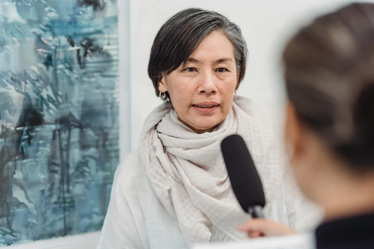 A woman with short gray hair and a white scarf is speaking into a microphone during an interview. A blurred figure holds the microphone.
