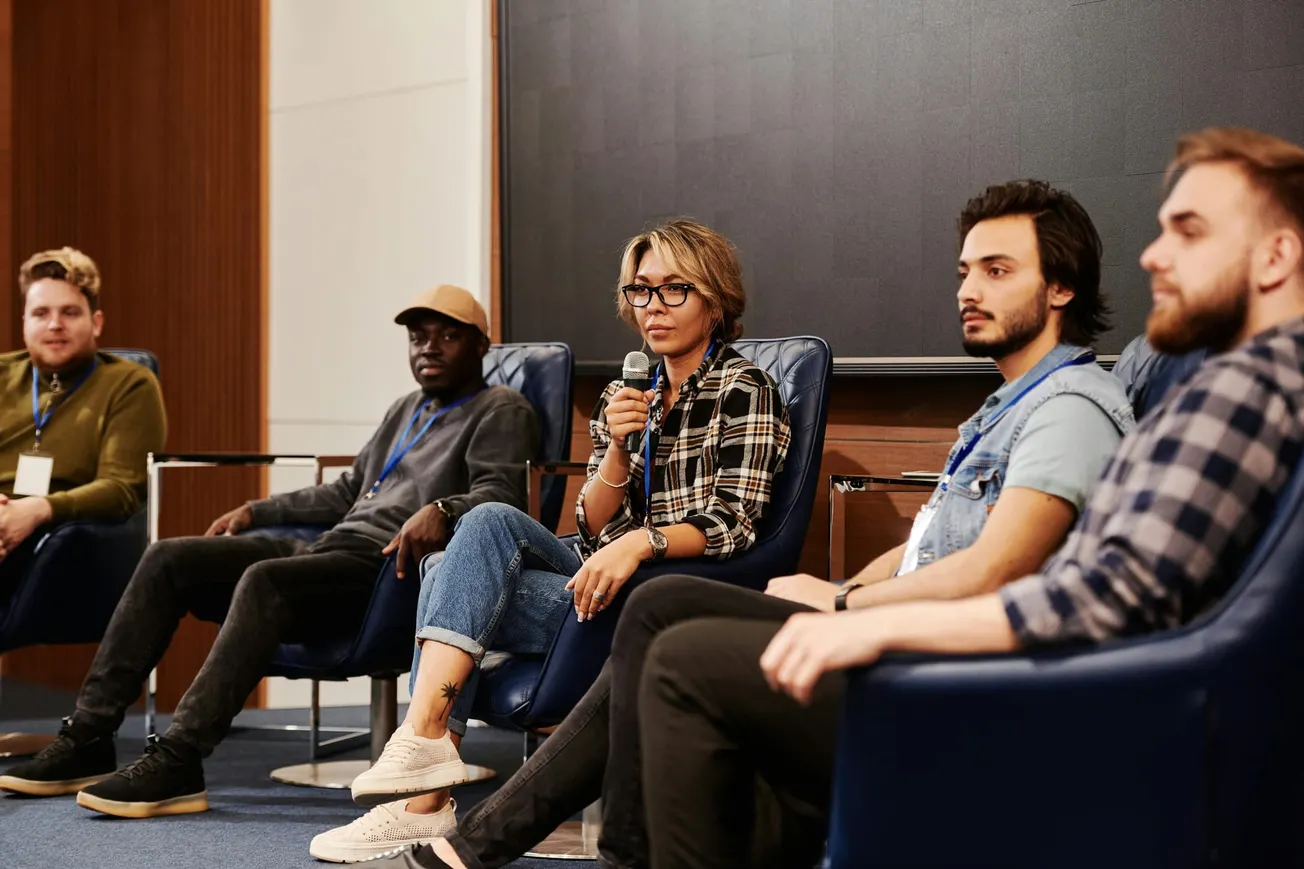 A diverse panel of five people seated on stage during a discussion. A woman with glasses holds a microphone, engaging the audience.