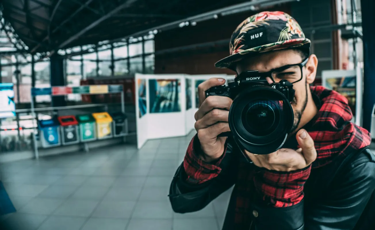Young man in a floral cap and red flannel focuses a camera in an indoor setting by colorful recycle bins.
